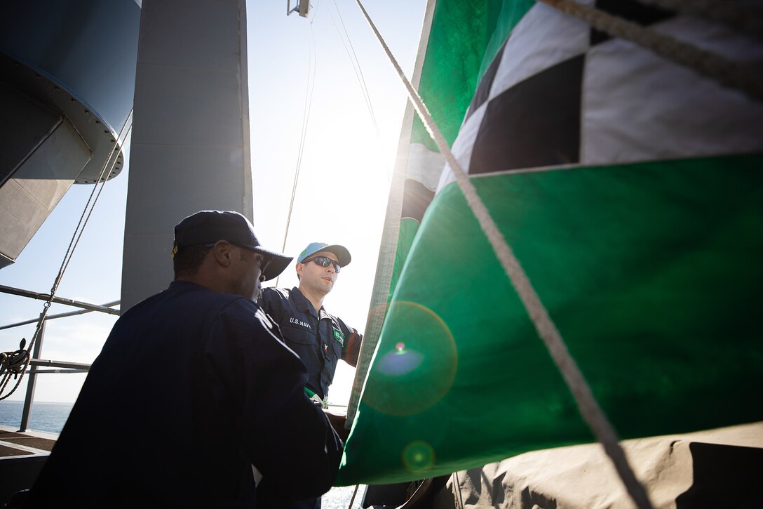 Two sailors work near a green flag with black and white squares on a ship's deck.