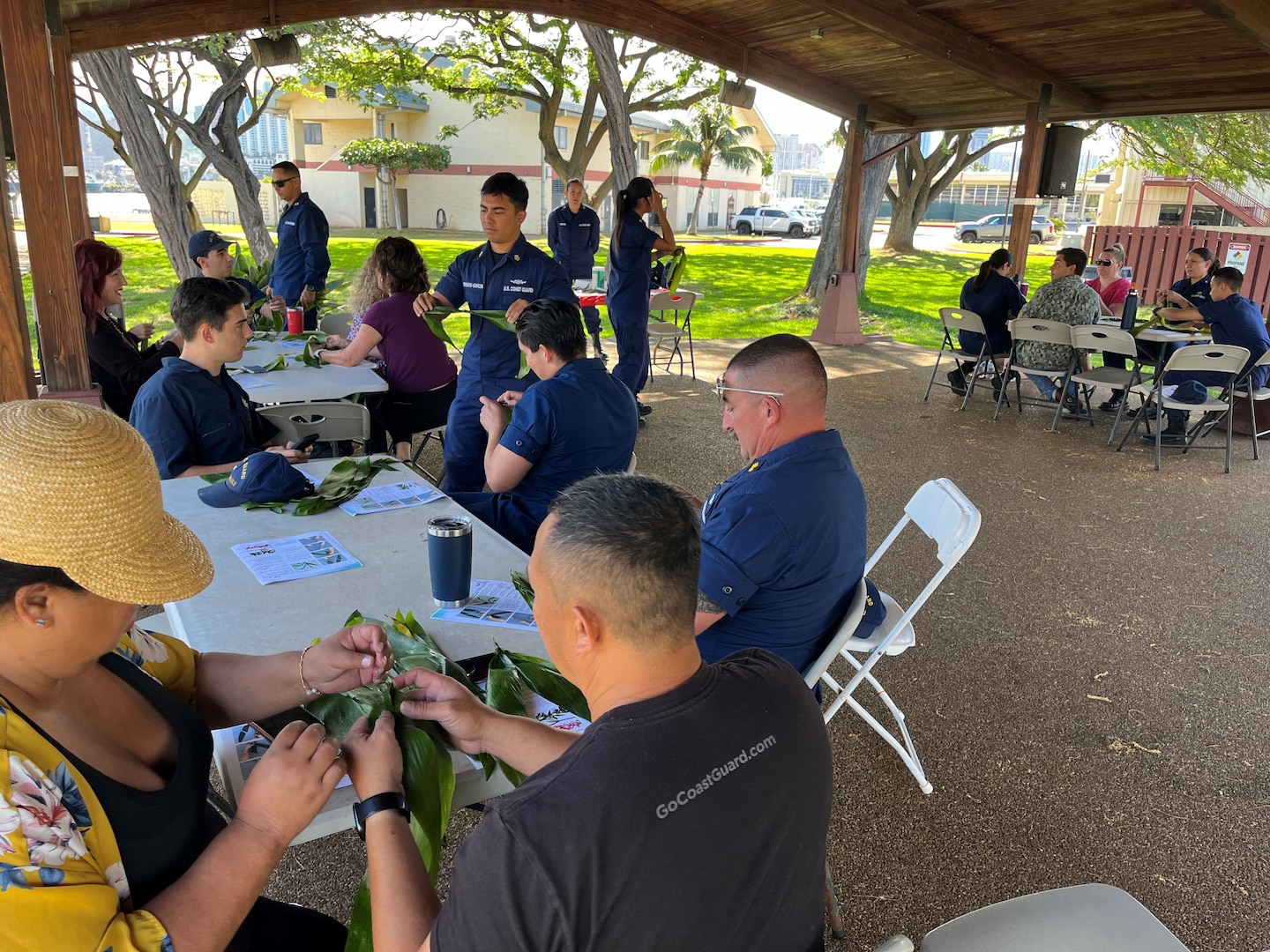 Coast Guard members and civilians sit outside in groups and are crafting leis.