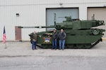 Three men wearing civilian clothes stand in front of a green combat vehicle