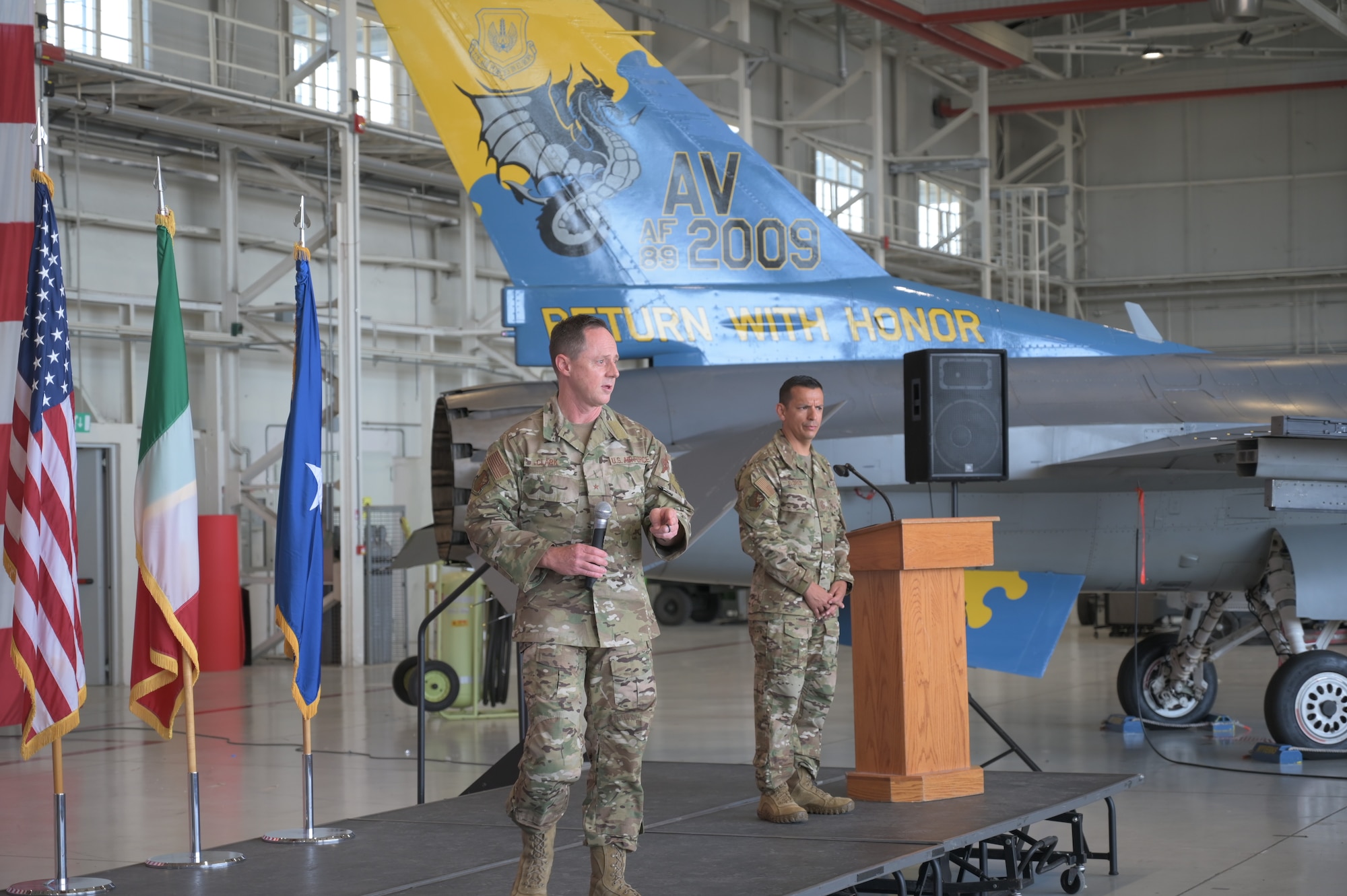 Two men speak in front of a flag and a jet with a painted tail that says, "Return with honor"
