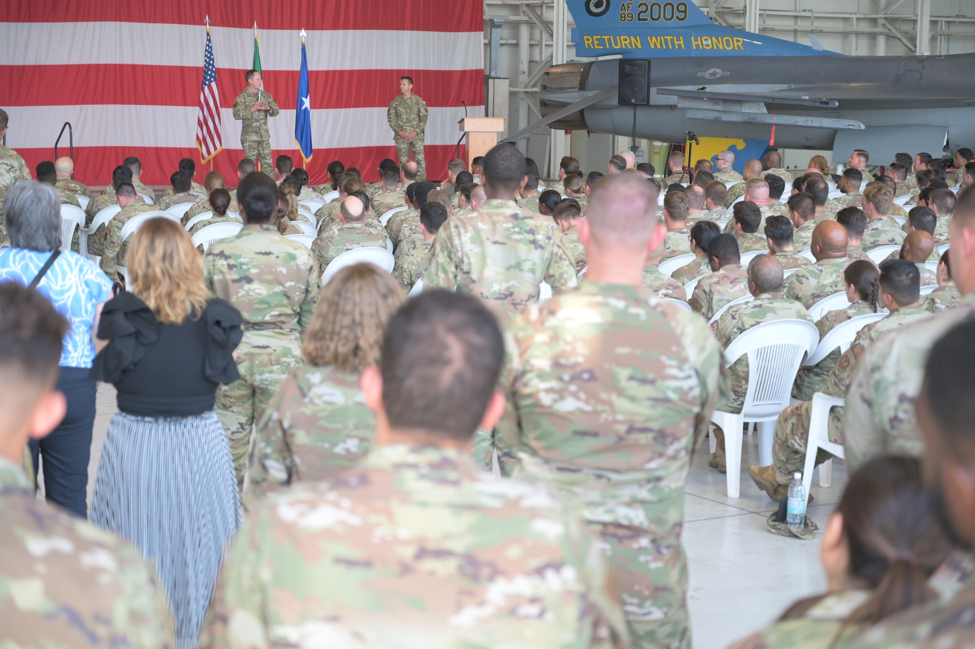 A crowd watches two men who are speaking in front of a large American flag.