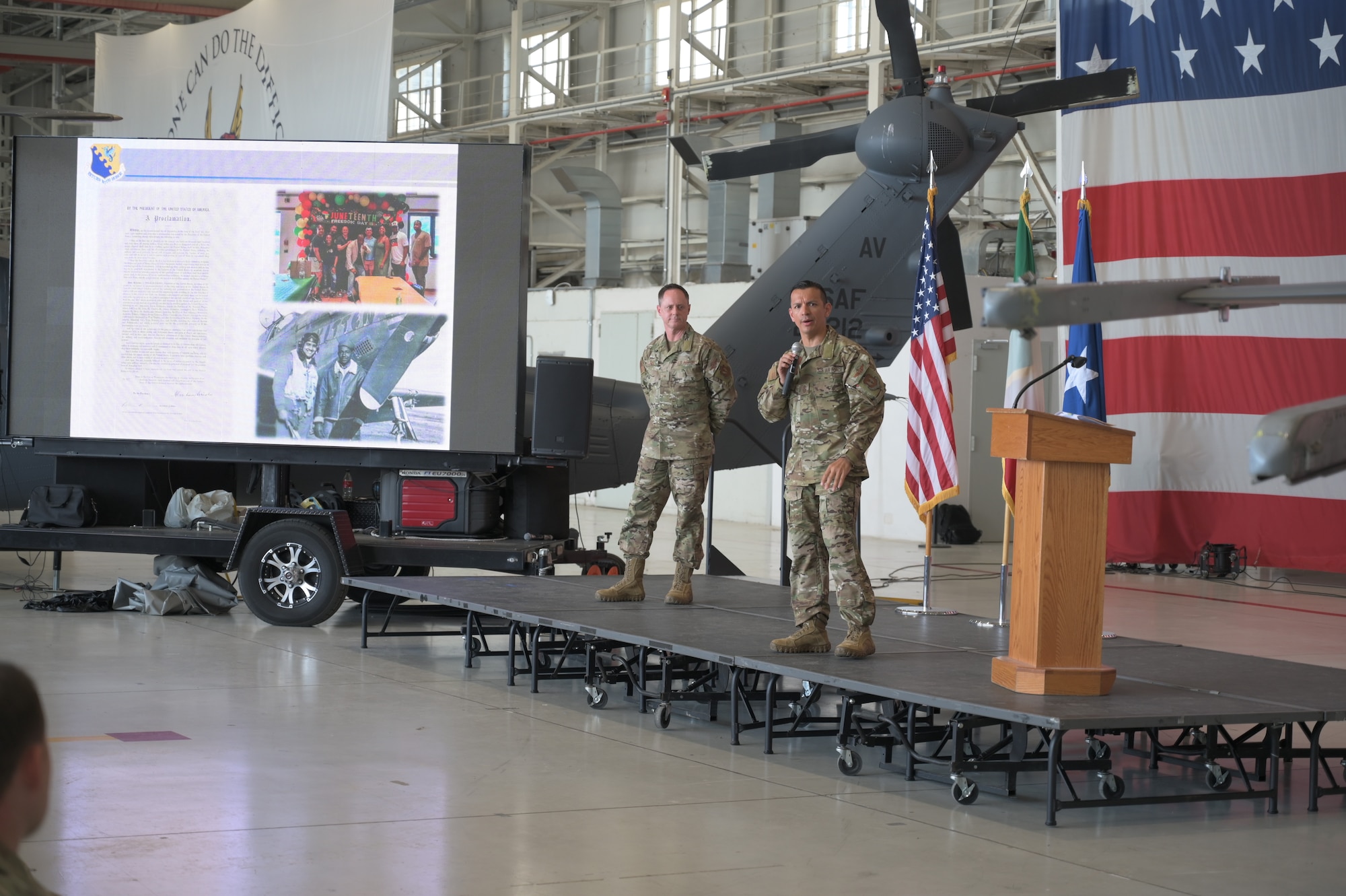 Two military members stand on a dais in front of a flag and large screen