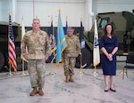 A woman wearing a blue dress and two men wearing Army uniforms stand in front of various flags
