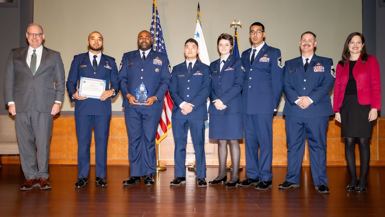 Eight people stand together on a stage. Six people are in military uniforms; the others are in civilian clothing.