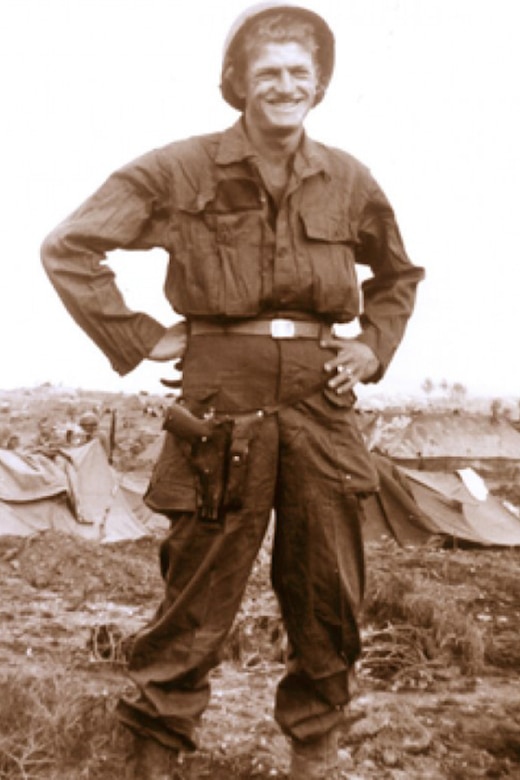 A smiling service member in combat uniform stands with their hands on their hips.