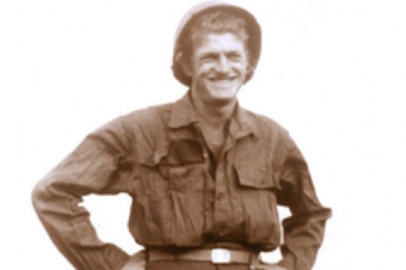 A smiling service member in combat uniform stands with their hands on their hips.