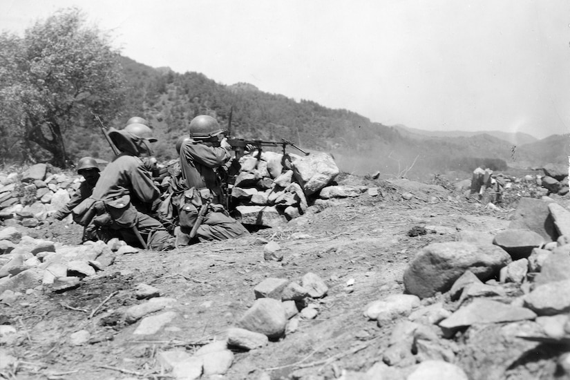 Two service members take cover behind a small barrier. One shoots a weapon into the distance.