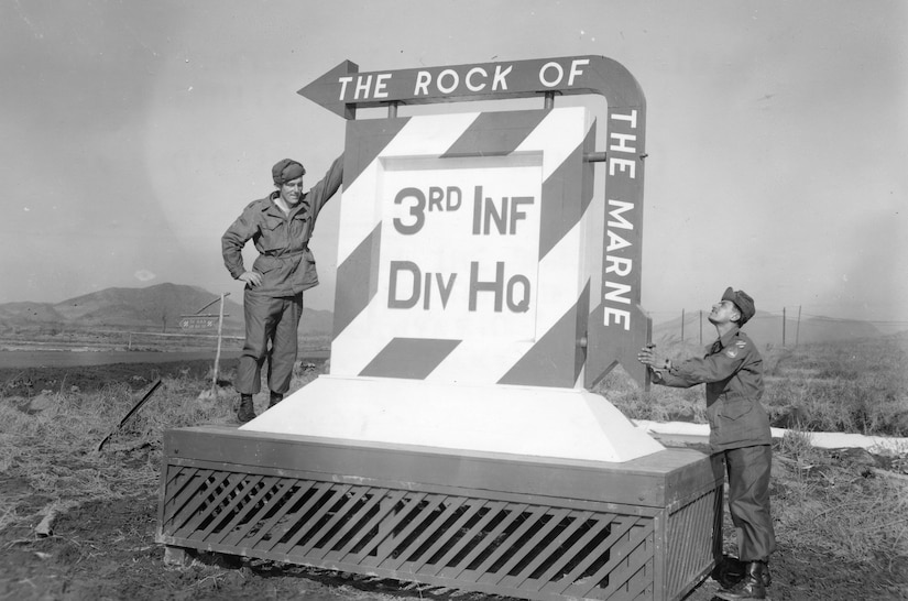 Two people stand beside a large road sign that reads “3rd Inf Div Hq.”