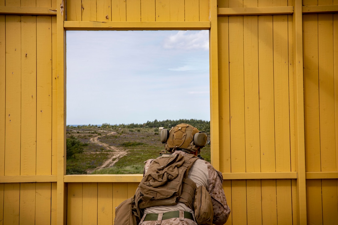 A Marine looks at grassy hills through an opening in a yellow wooden structure.