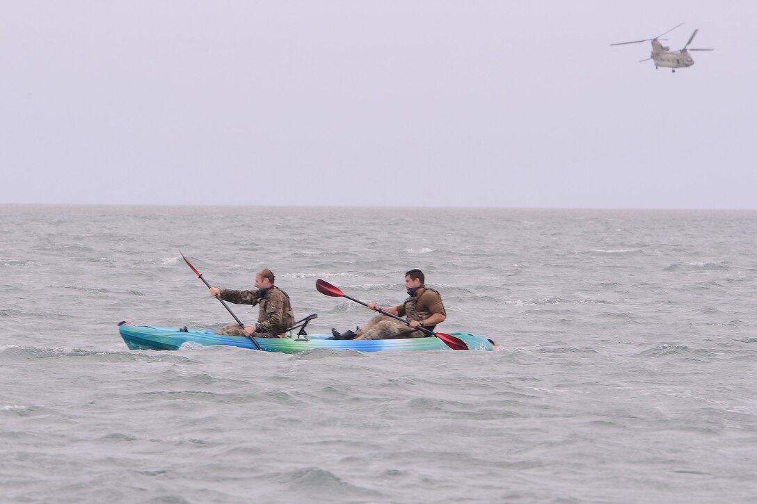 Two soldiers use oars in a kayak in a body of water as a helicopter flies above to the right in the background.