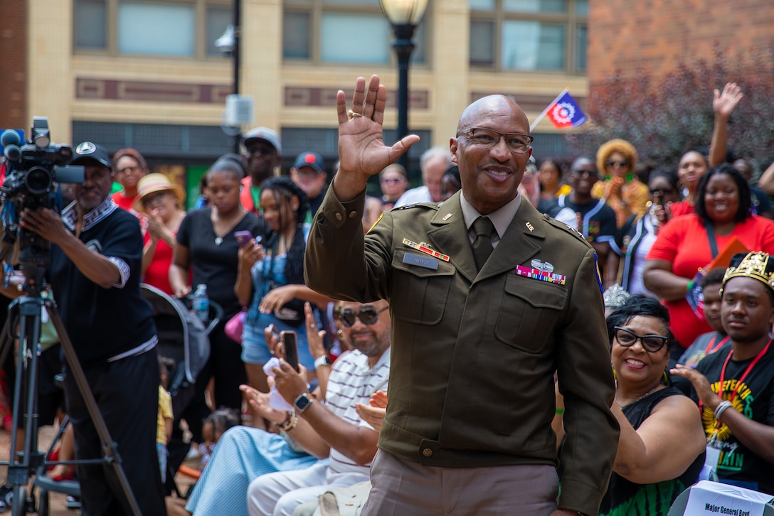 A soldier smiles and waves as a seated and standing crowd cheers from the background while a cameraperson records.