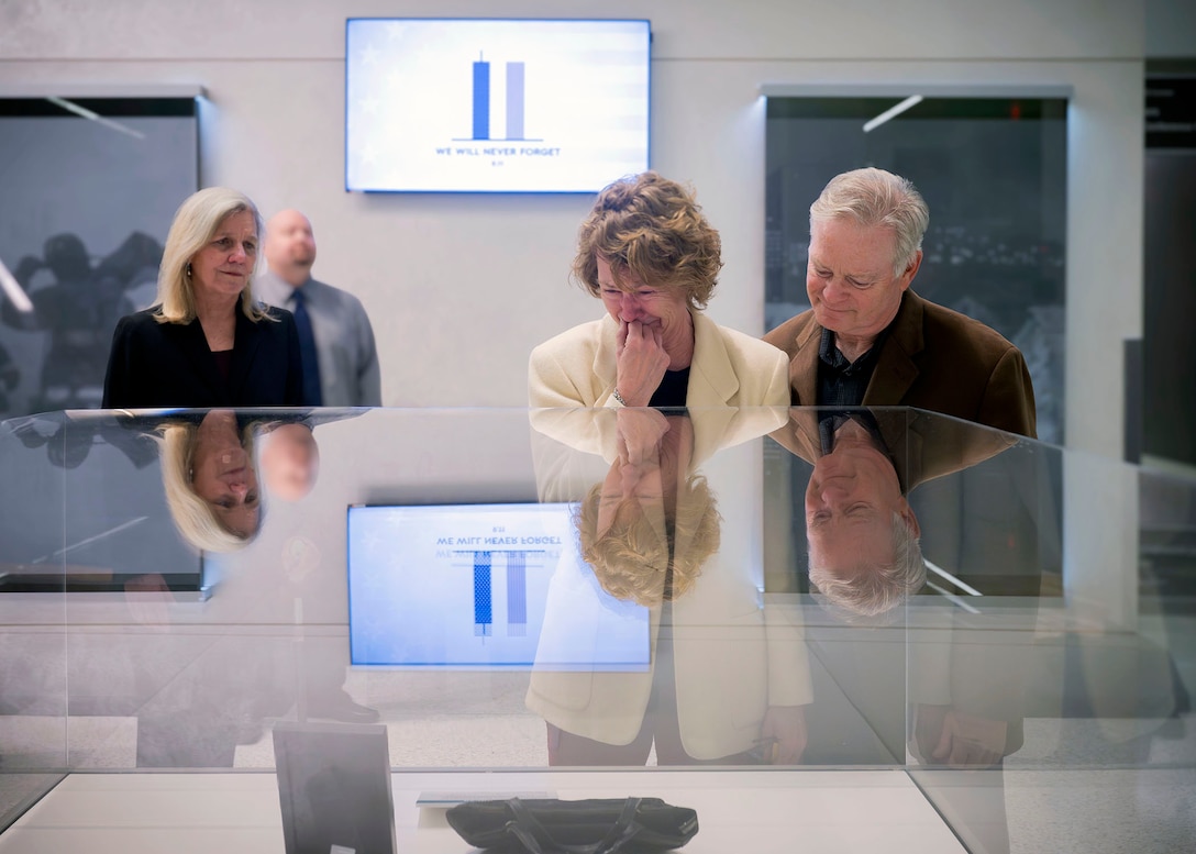 Deora Bodley's mother Debby, a women in a cream colored jacket, cries as she views her daughter's display in NSA's 9/11 Memorial. She is comforted by her partner, a man wearing a brown jacket. Deputy Wendy Noble, in a black suit, looks on.
