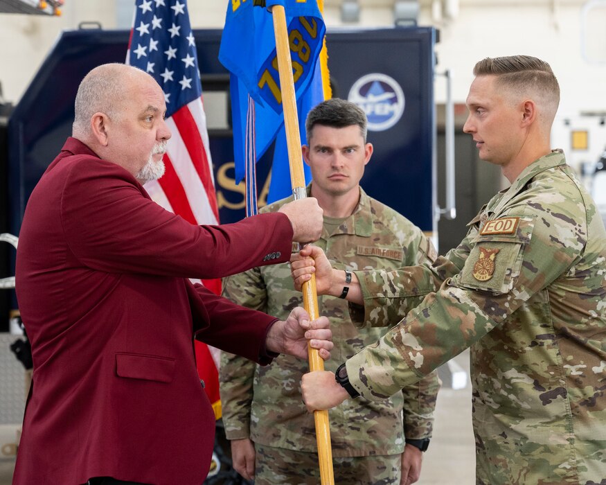 A man in a red blazer stands on the left facing a man in camouflage uniform. They hold a unit flag between them.
