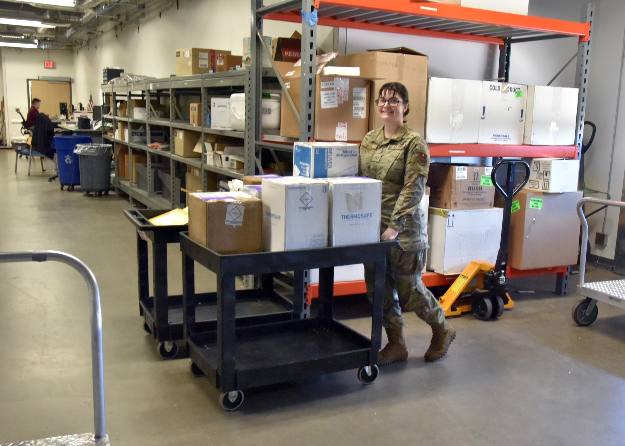 An Airman pushes a cart with boxes on it in a warehouse.