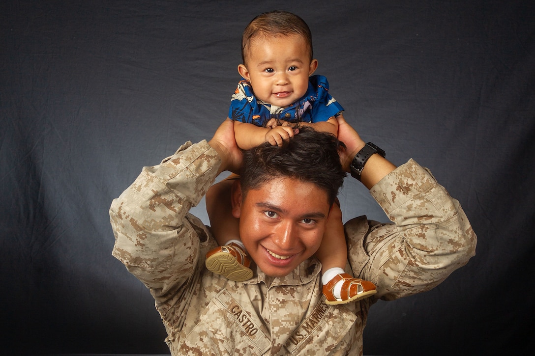 A Marine smiles while holding a toddler on his shoulders in front of a dark background.