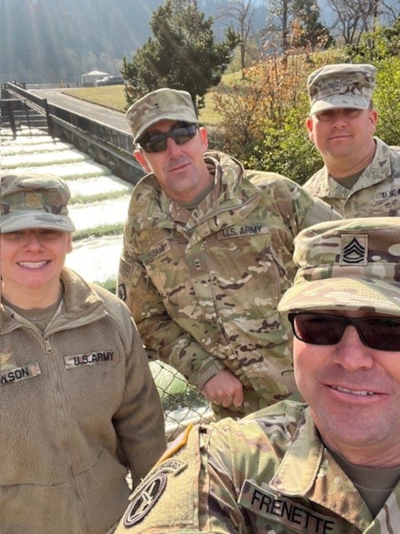 Four soldiers in uniform stand with trees and a concrete path in the background.