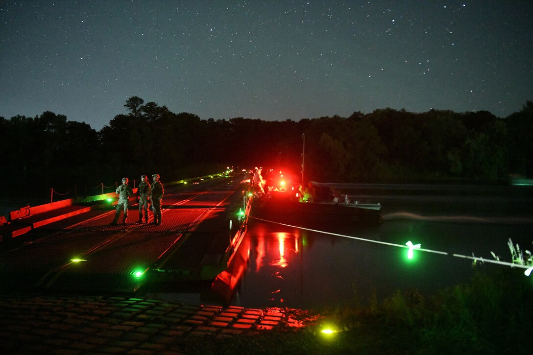 Service members unload supplies from a small boat to assemble a bridge at night. Green and red lights illuminate the scene.