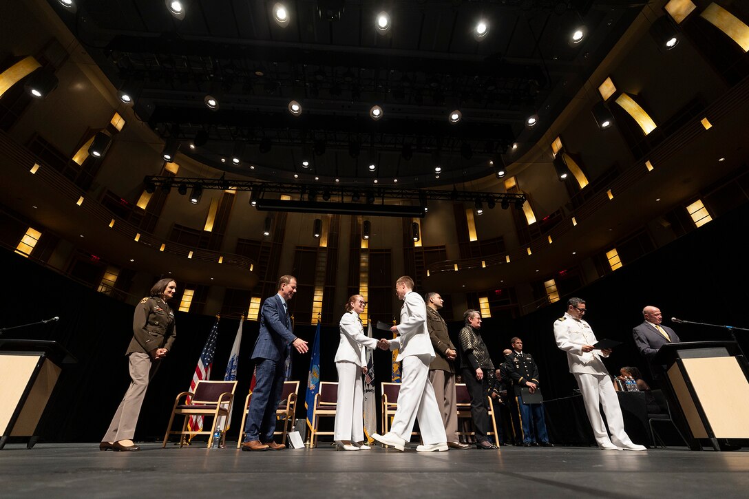 A graduate walks across the stage and shakes hands with a uniformed service member as others watch.