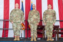 Three men in military uniform stand at attention on a stage with an American flag draped behind them.