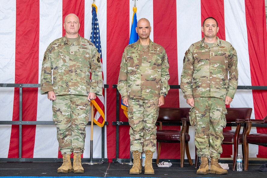 Three men in military uniform stand at attention on a stage with an American flag draped behind them.