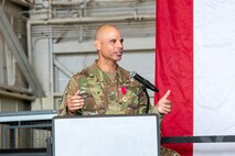 A man in military uniform speaks at a podium while gesturing towards an out-of-frame crowd.