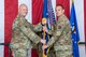 Two men in military uniforms simultaneously grasp a flag pole on a stage.