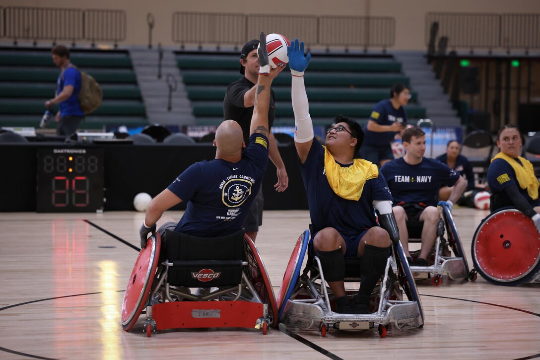 Two Navy athletes using wheelchairs meet in the center for tipoff during a rugby game on a basketball court with empty bleachers in the background.