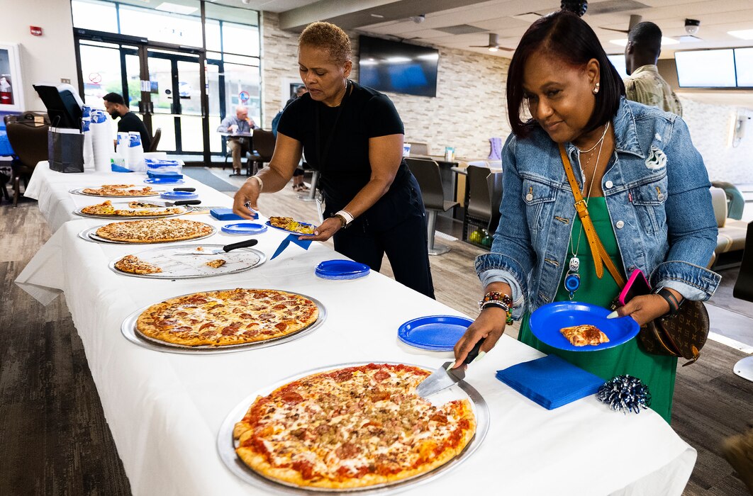 A woman serves herself a slice of pizza from a long table of pies. Another woman serves herself in the background.