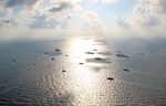 Twenty military ships at sea sit on or nearby the sun's reflection off the ocean's surface on clear day.