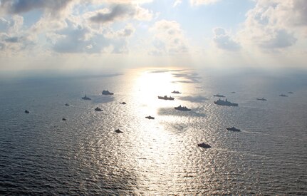 Twenty military ships at sea sit on or nearby the sun's reflection off the ocean's surface on clear day.