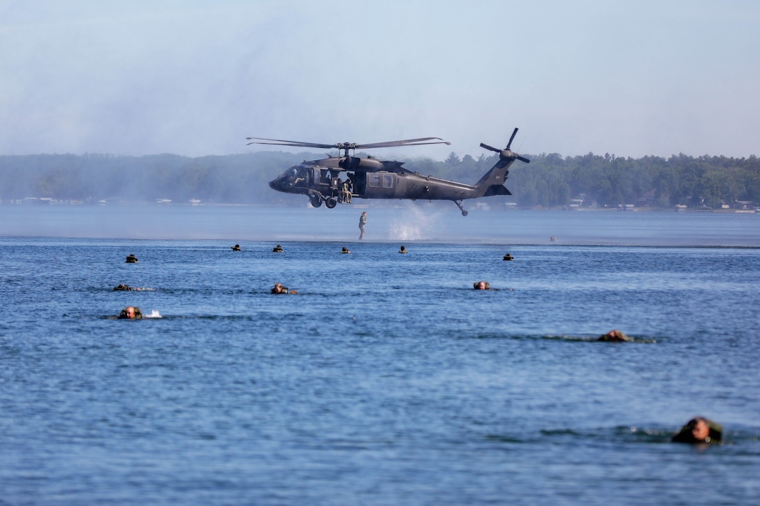 A soldier jumps from a low-flying helicopter into the water as other soldiers float nearby.