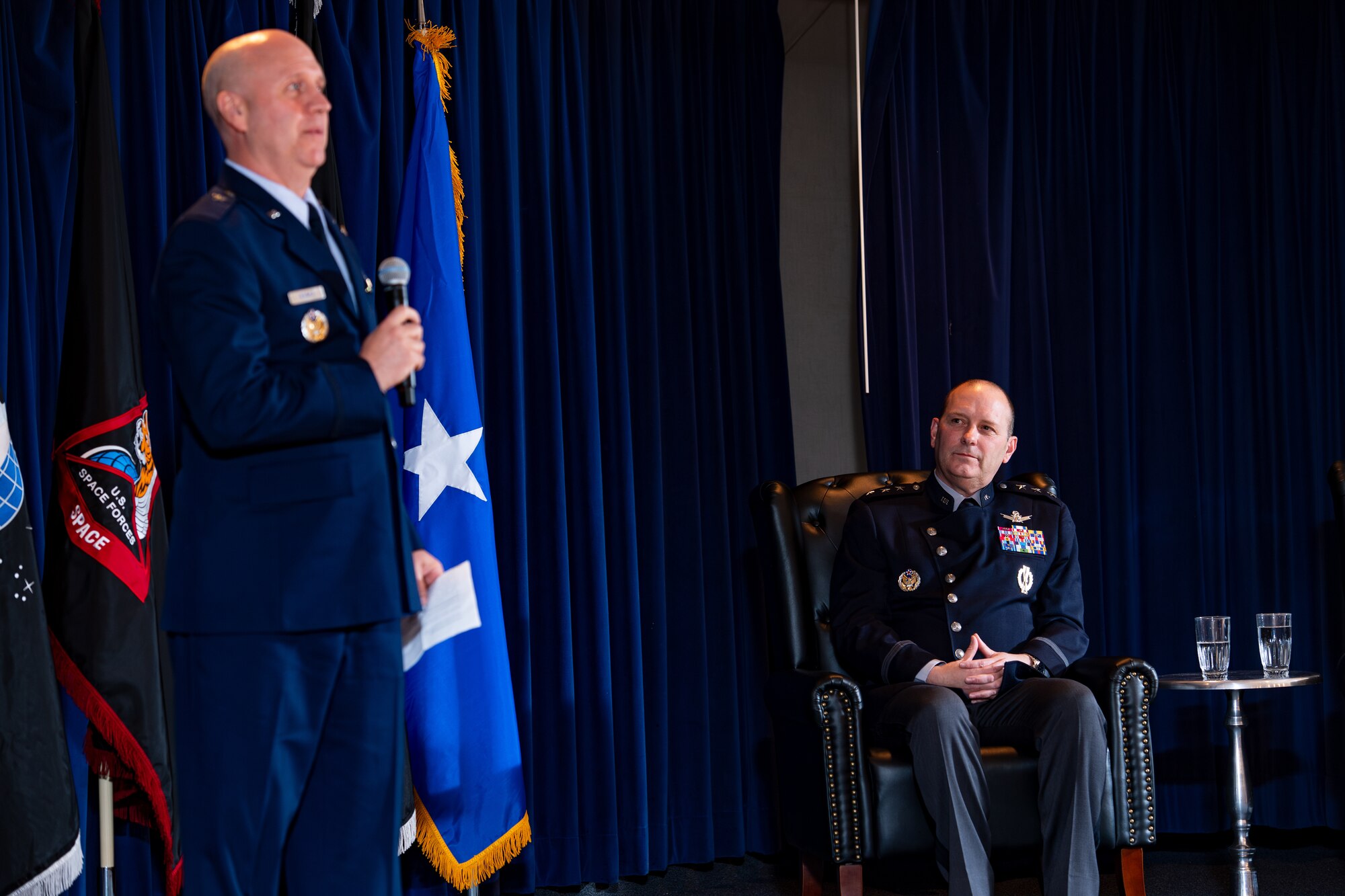 Man in uniform sits in chair listening to man in uniform holding microphone