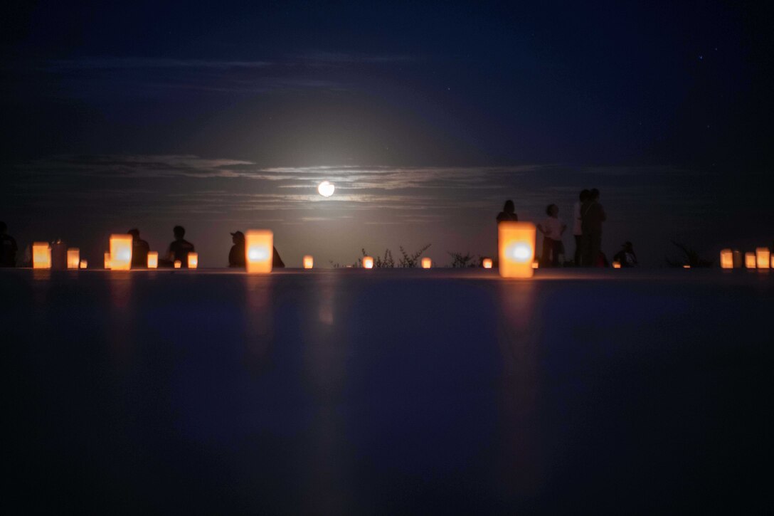 Lit candles lay in the foreground as U.S. sailors, civilians and Japanese volunteers stand in the background under a full moon.