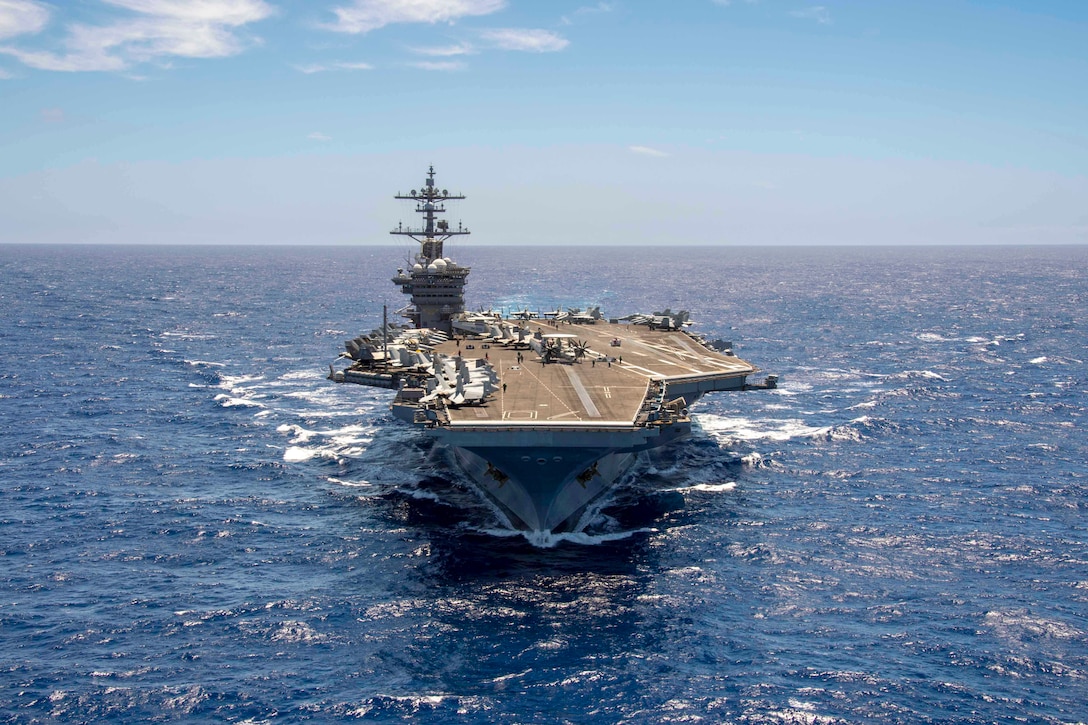 Sailors work near dozens of aircraft on the flight deck of a large ship transiting a body of water.