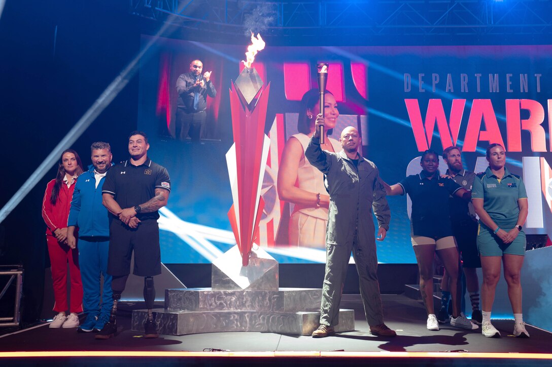 Athletes stand in formation on a stage next to a life-size lit cauldron with a monitor behind them.