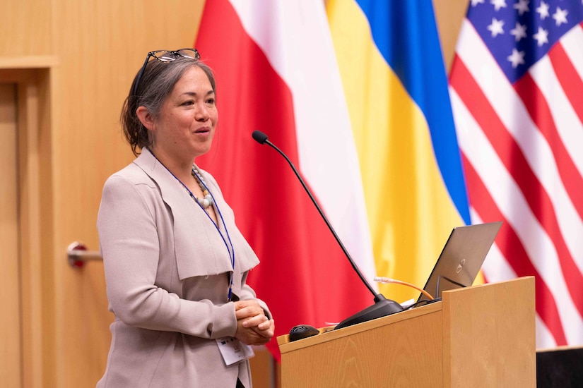 A woman wearing business attire stands behind a lectern with a microphone with several flags in the background.