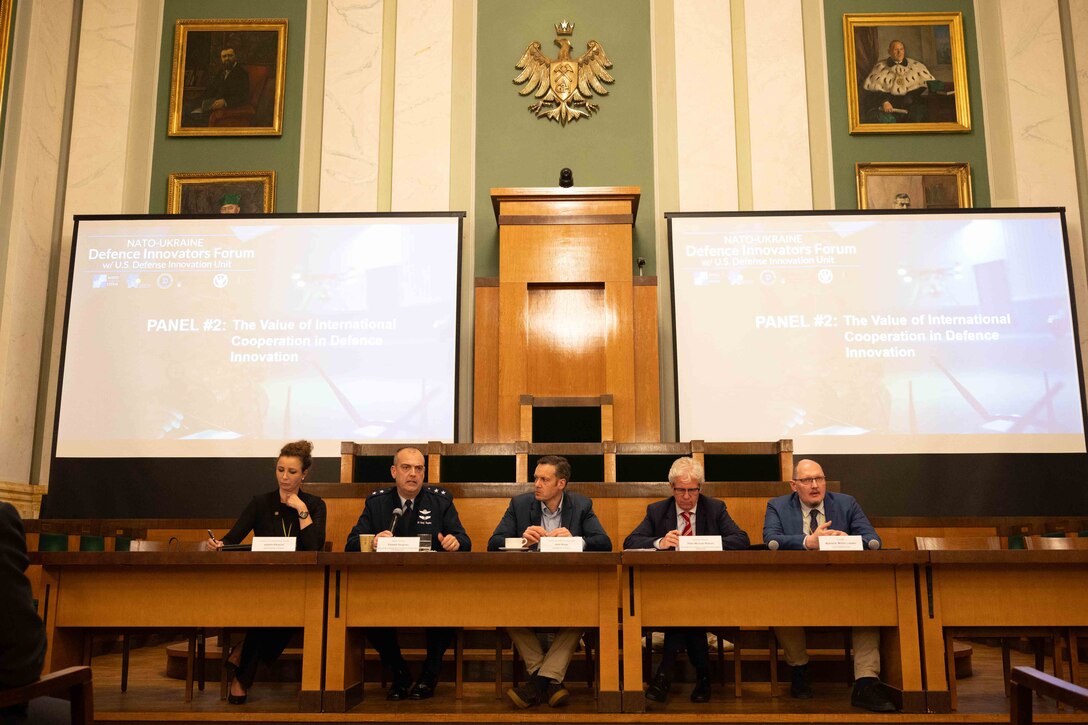 Five people, including one military service member, sit shoulder-to-shoulder at a table with two presentation screens behind them.