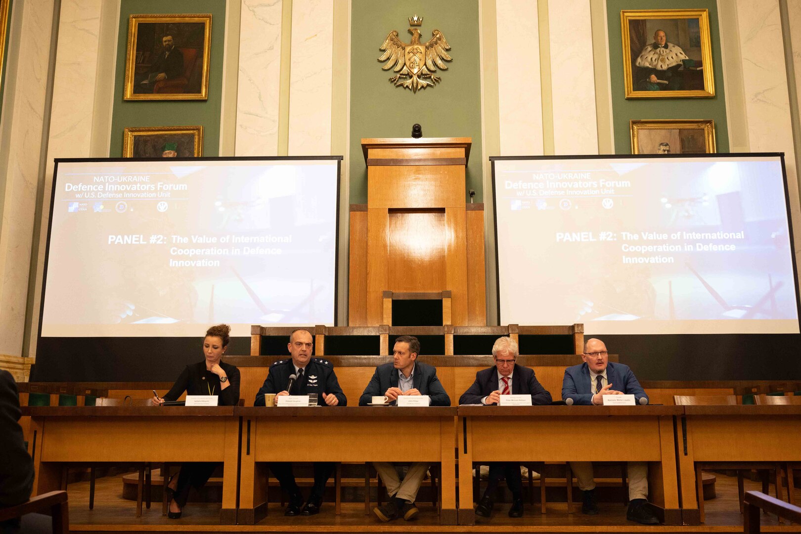 Five people, including one military service member, sit shoulder-to-shoulder at a table with two presentation screens behind them.