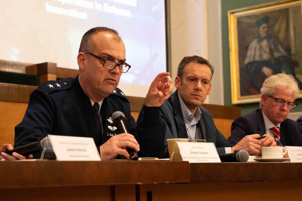 A military service member and two men wearing business suits sit at a table with microphones.
