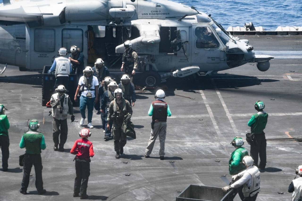 A group of people mill around outside of an aircraft on the deck of a carrier.