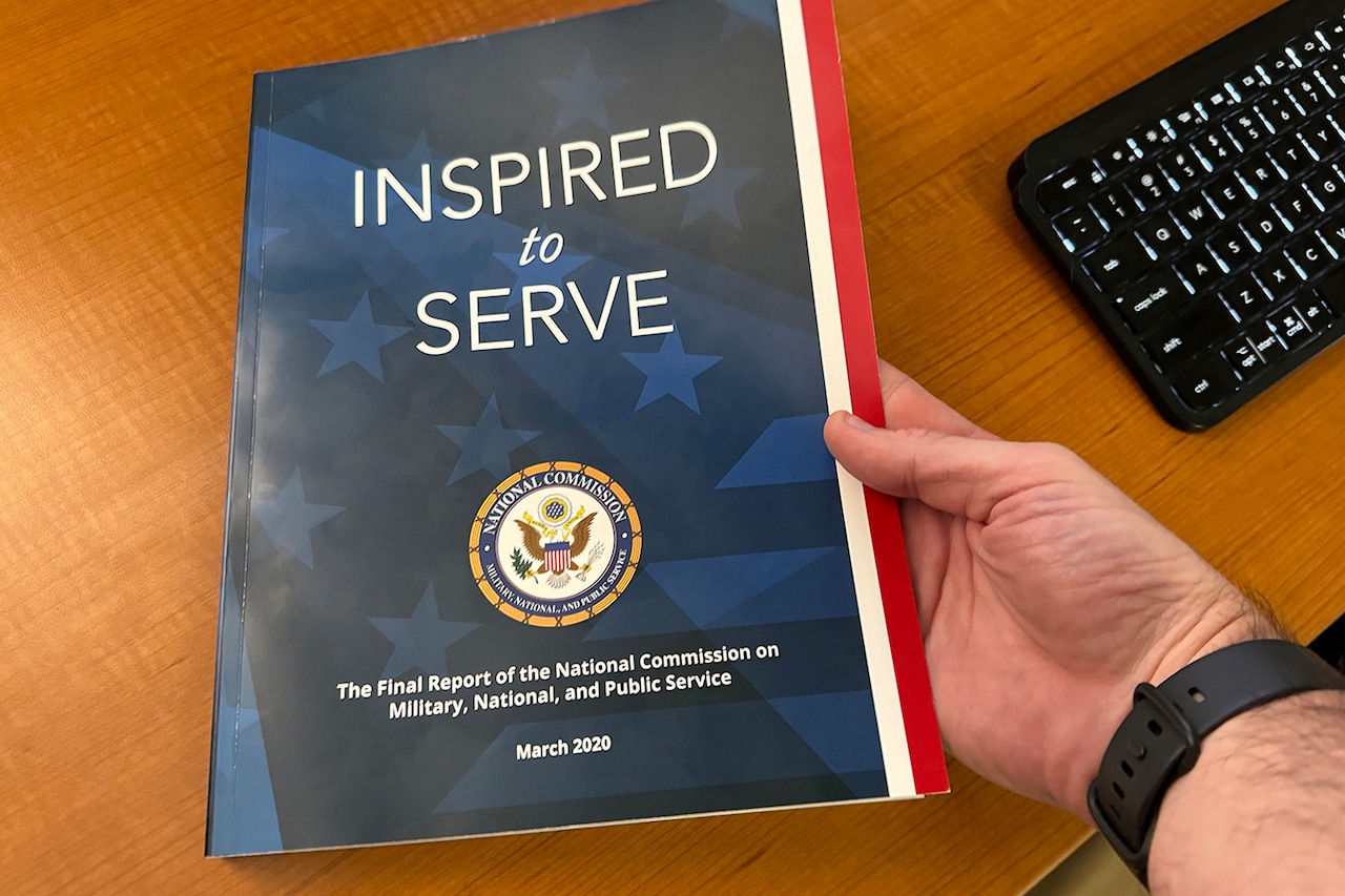 A hand holds an 8-1/2” x 11” soft-cover book atop a brown wooden desktop. The book is titled "Inspired to Serve: The Final Report of the National Commission on Military, National, and Public Service."