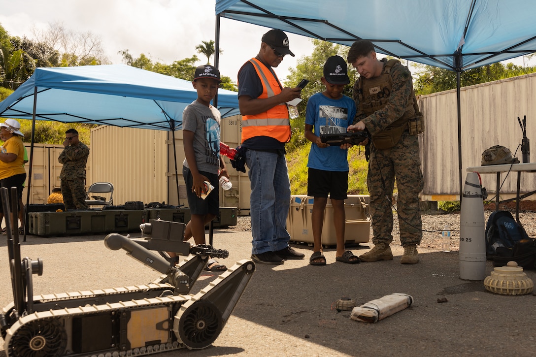 U.S. Marine Corps Sgt. Adam Towle, an explosive ordnance technician with Combat Logistics Battalion 13, Combat Logistics Regiment 17, 1st Marine Logistics Group, and a native of South Kingstown, Rhode Island, shows local visitors a small unmanned ground vehicle during a military equipment exposition as part of exercise Valiant Shield 24 at Palau International Airport, Airai, Palau, June 13, 2024. U.S. joint forces conducting exercise Valiant Shield 24 held a military equipment exposition to offer insight into military operations and build relationships with the Palauan community. I Marine Expeditionary Force conducts exercises like Valiant Shield to enhance interoperability with allied and partner forces and expand the operational capability of the Marine Air-Ground Task Force in the Indo-Pacific region. (U.S. Marine Corps photo by Lance Cpl. Hannah Hollerud)