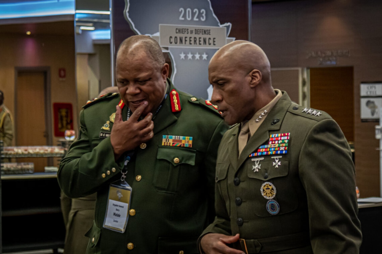 Two men in military uniforms stand near each other and speak.