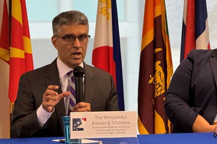 A person wearing a business suit and glasses is sitting at a table while speaking into a microphone.