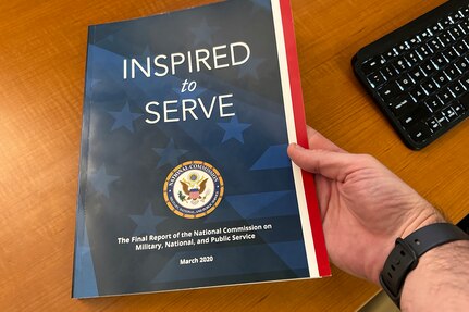 A hand holds an 8-1/2” x 11” soft-cover book atop a brown wooden desktop. The book is titled "Inspired to Serve: The Final Report of the National Commission on Military, National, and Public Service."