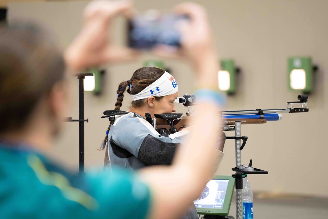 An airman aims a rifle while a spectator in the foreground holds a camera to take a picture.