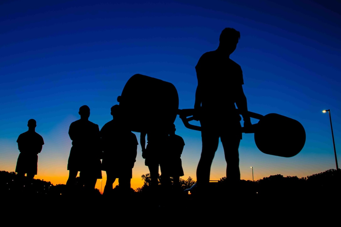 A group of soldiers are silhouetted against a twilight sky. One soldier lifts a heavy free weight while others watch.