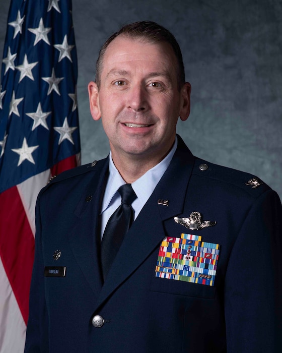 A man in an Air Force dress blues uniform stands in front of an American Flag and a grey background.