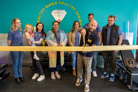 A group of people pose behind a celebratory golden ribbon. A man poses cutting the ribbon with a large pair of scissors.