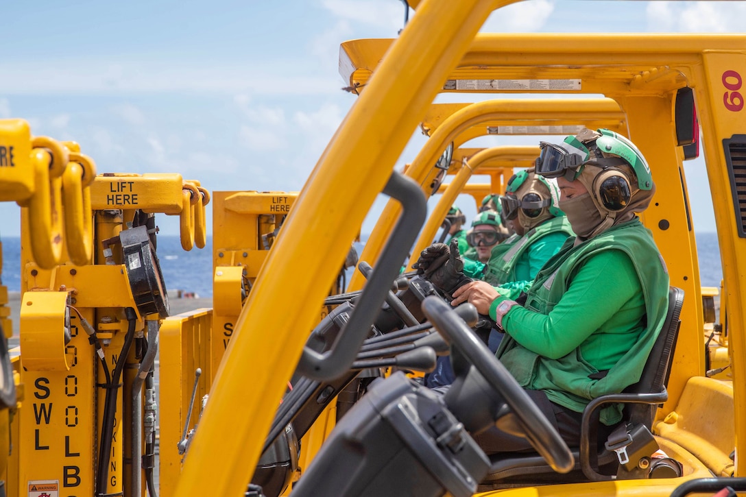 Five sailors sit in a row in orangish yellow fuel vehicles aboard a ship at sea.